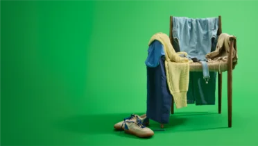 A wooden chair draped with various clothes, including a blue shirt and yellow sweater, with shoes on the green floor.